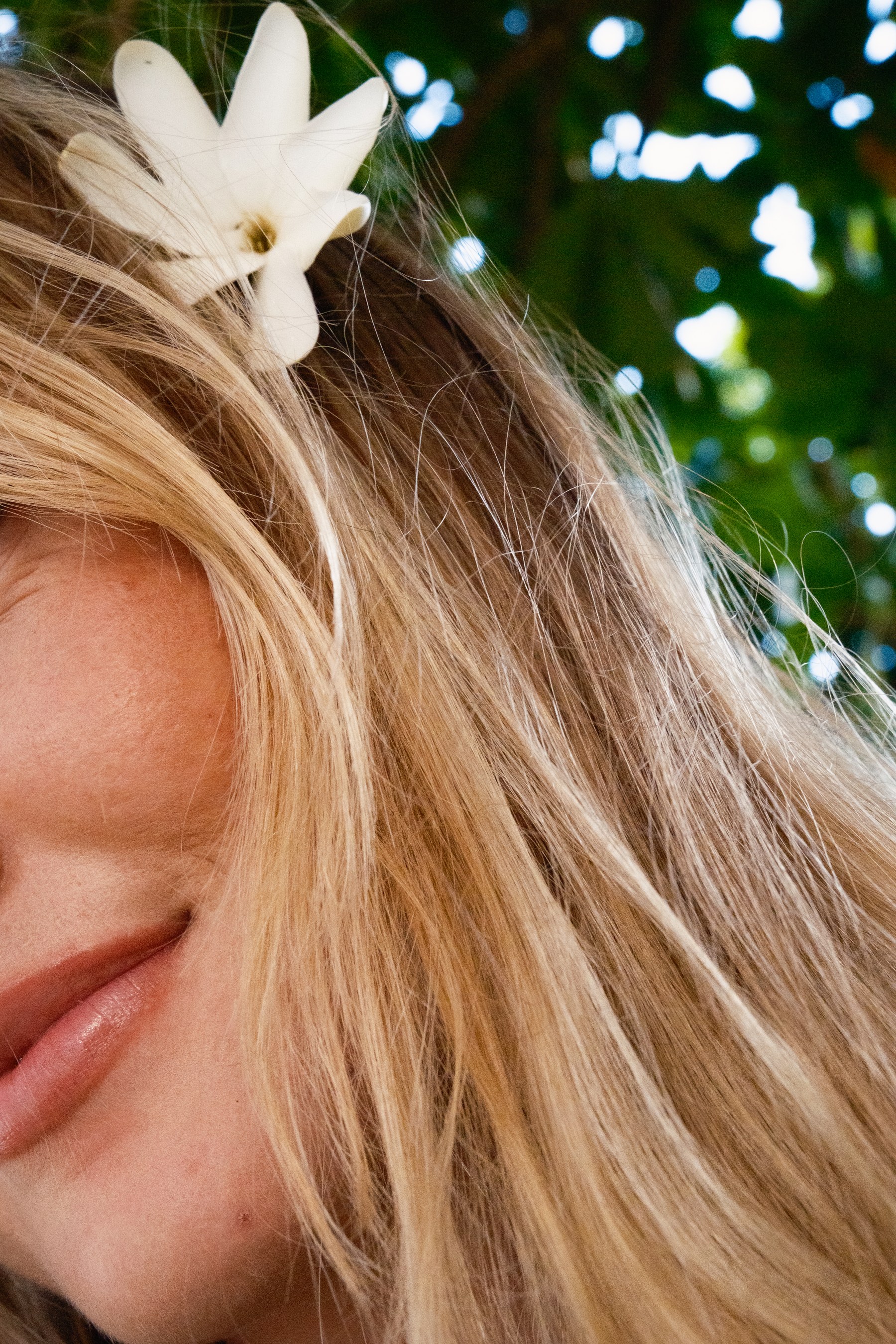 Close-up portrait with a white flower tucked into blonde hair against green foliage